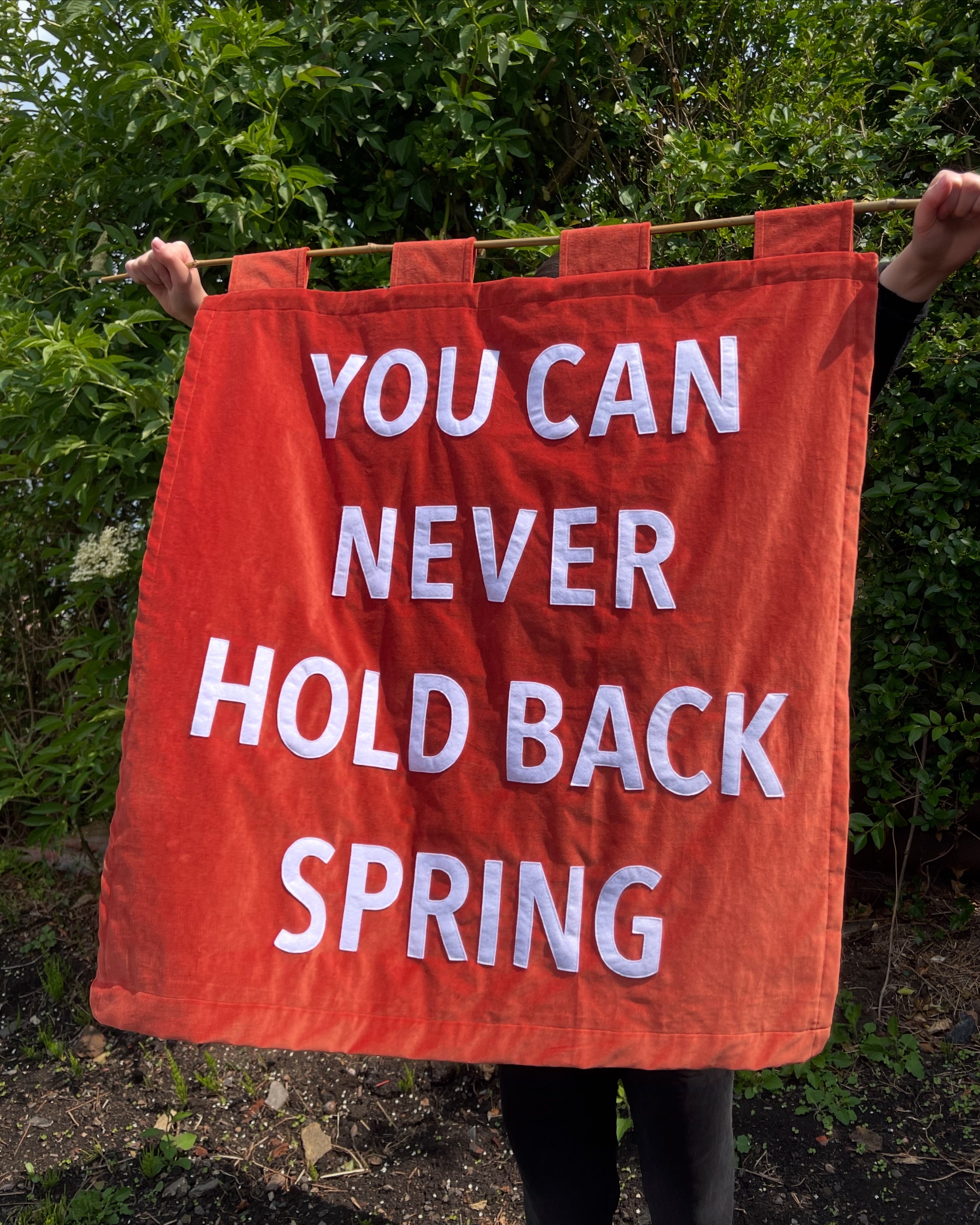 Orange velvet banner with white text 'YOU CAN NEVER HOLD BACK SPRING' held by a person outdoors.