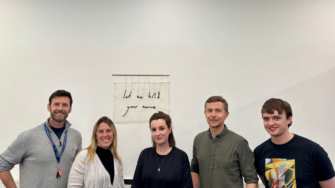 A group of five people stand in front of a textile artwork which reads "let me hold your nerve"