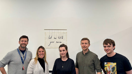 A group of five people stand in front of a textile artwork which reads "let me hold your nerve"