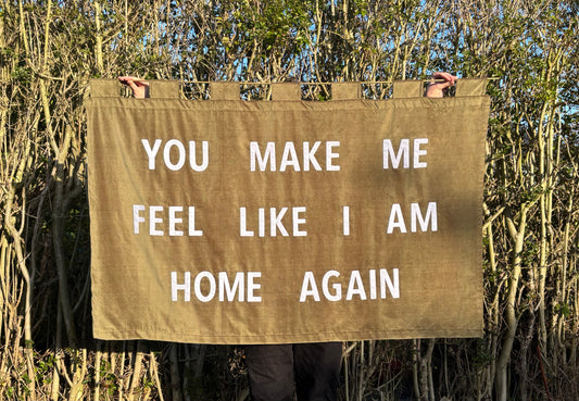 Person holding a large olive green velvet banner which reads "YOU MAKE ME FEEL LIKE I AM HOME AGAIN" in front of trees