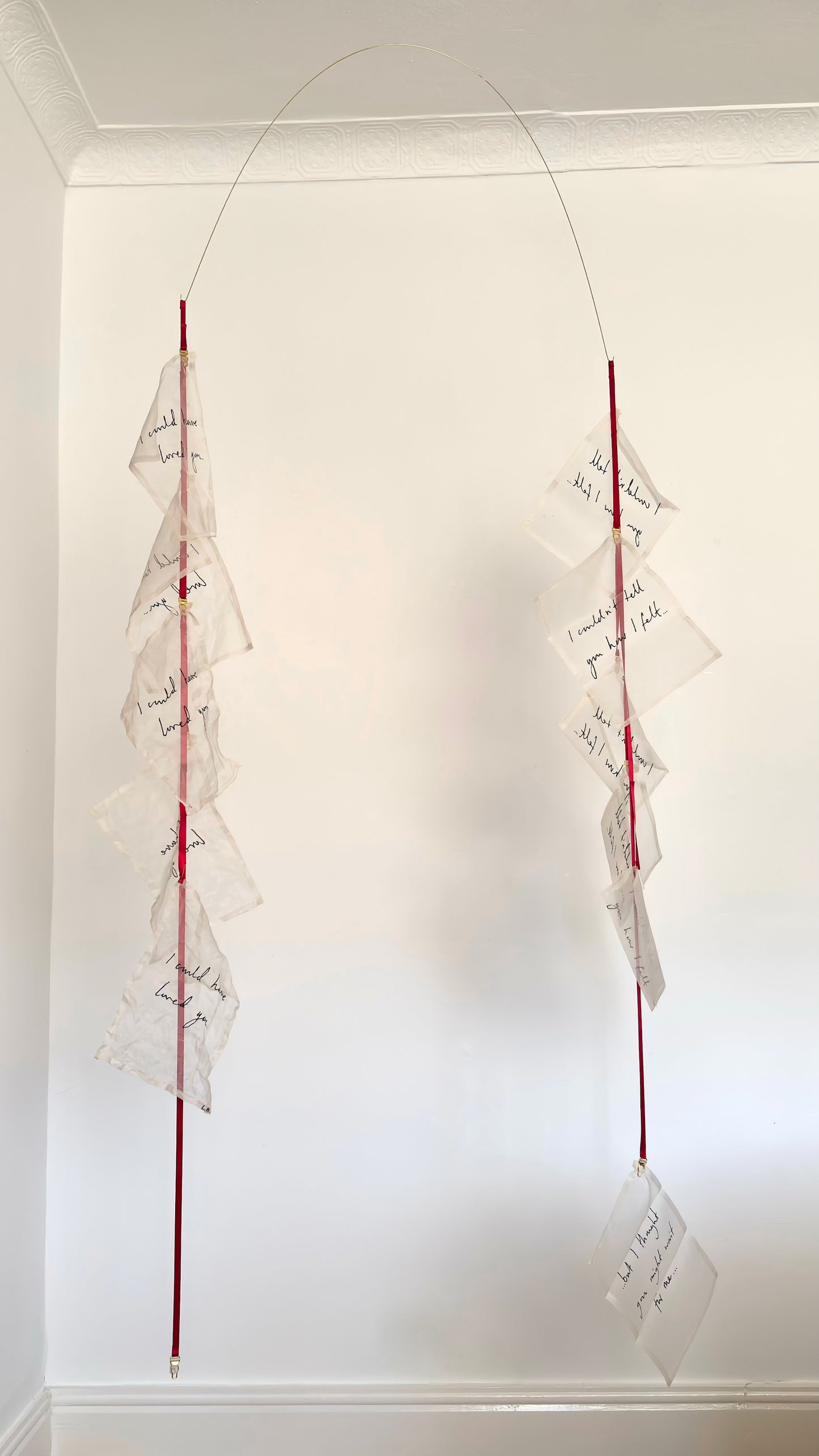 A kinetic textile sculpture, a brass arc with silk handwritten letters suspended from red ribbon.