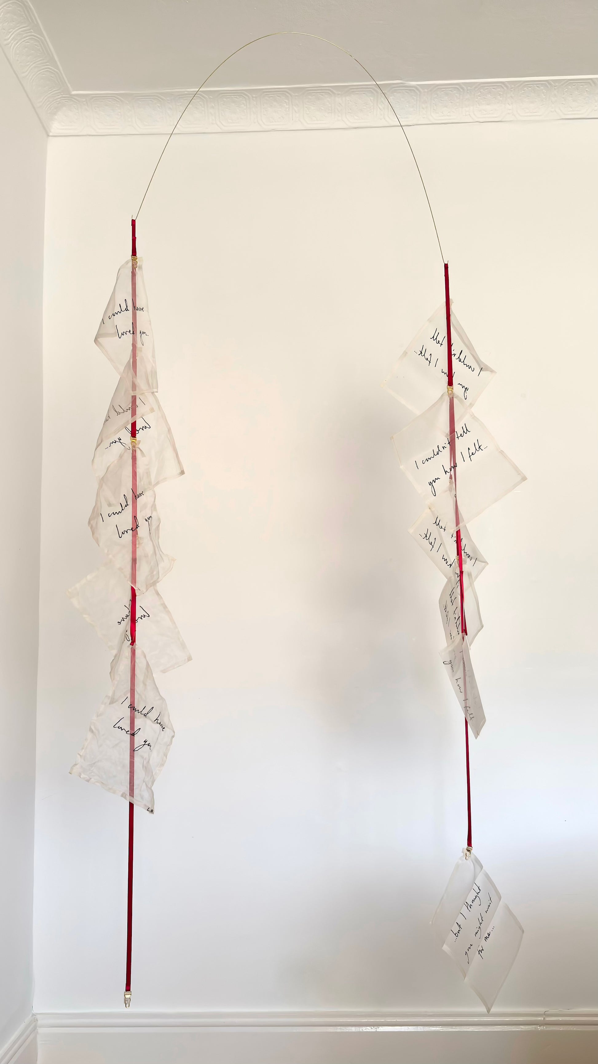 A kinetic textile sculpture, a brass arc with silk handwritten letters suspended from red ribbon.