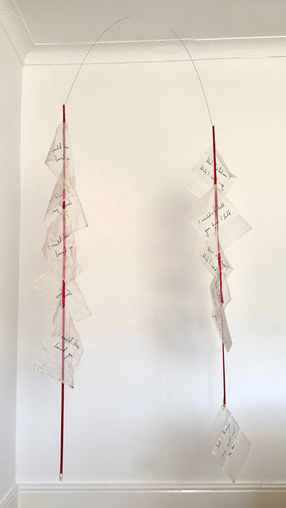 A kinetic textile sculpture, a brass arc with silk handwritten letters suspended from red ribbon.