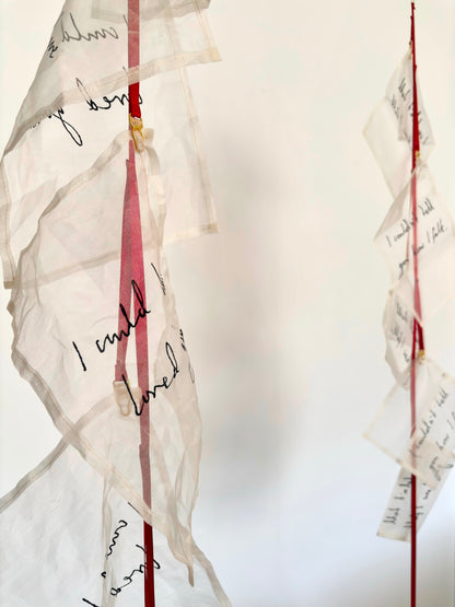 An abstract textile sculpture with multiple translucent silk handwritten letters attached to red ribbon, and suspender clasps, photographed against a plain light background.