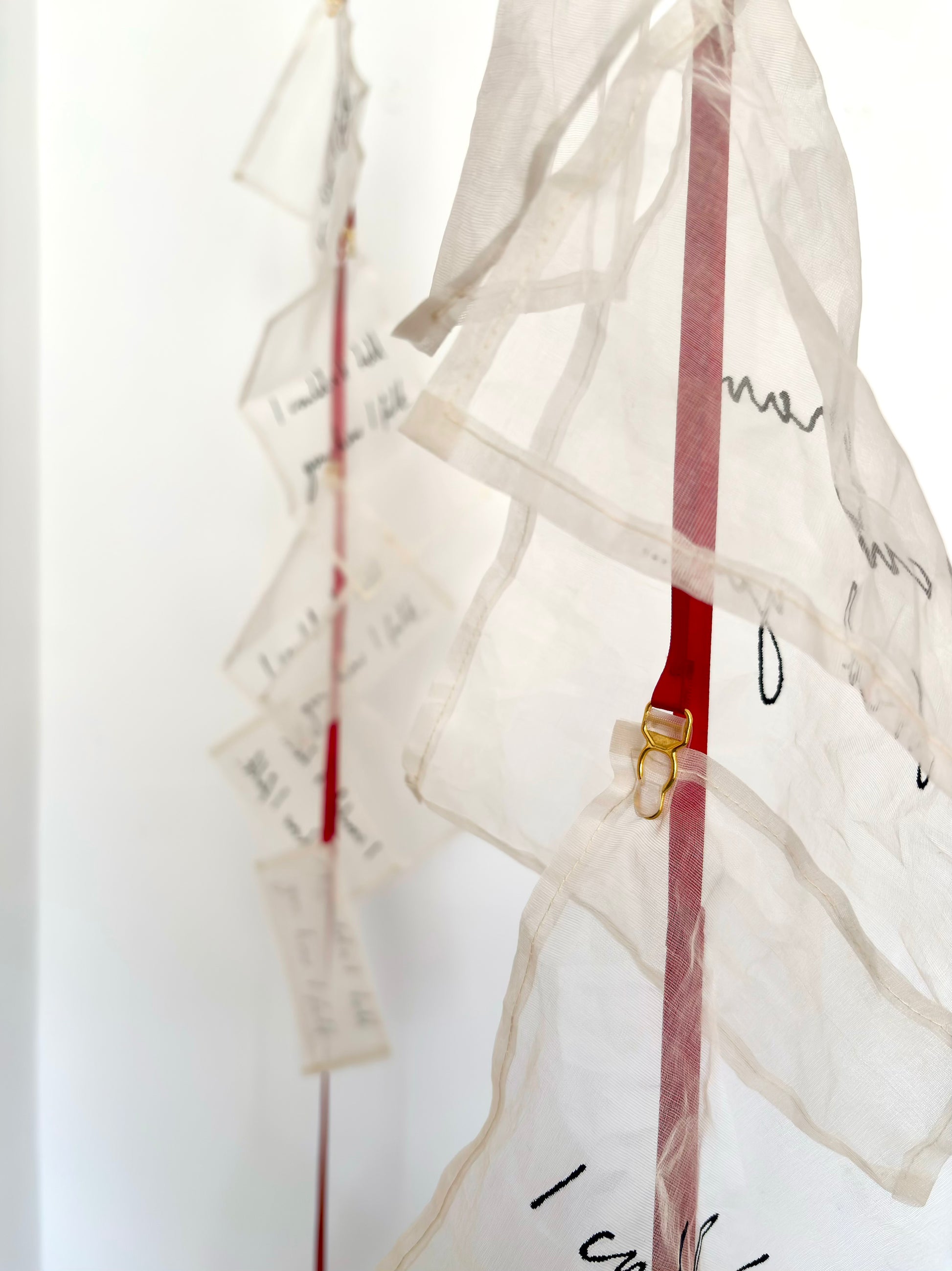 An abstract textile sculpture with multiple translucent silk handwritten letters attached to red ribbon, and suspender clasps, photographed against a plain light background.