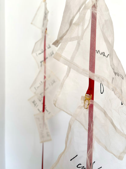 An abstract textile sculpture with multiple translucent silk handwritten letters attached to red ribbon, and suspender clasps, photographed against a plain light background.