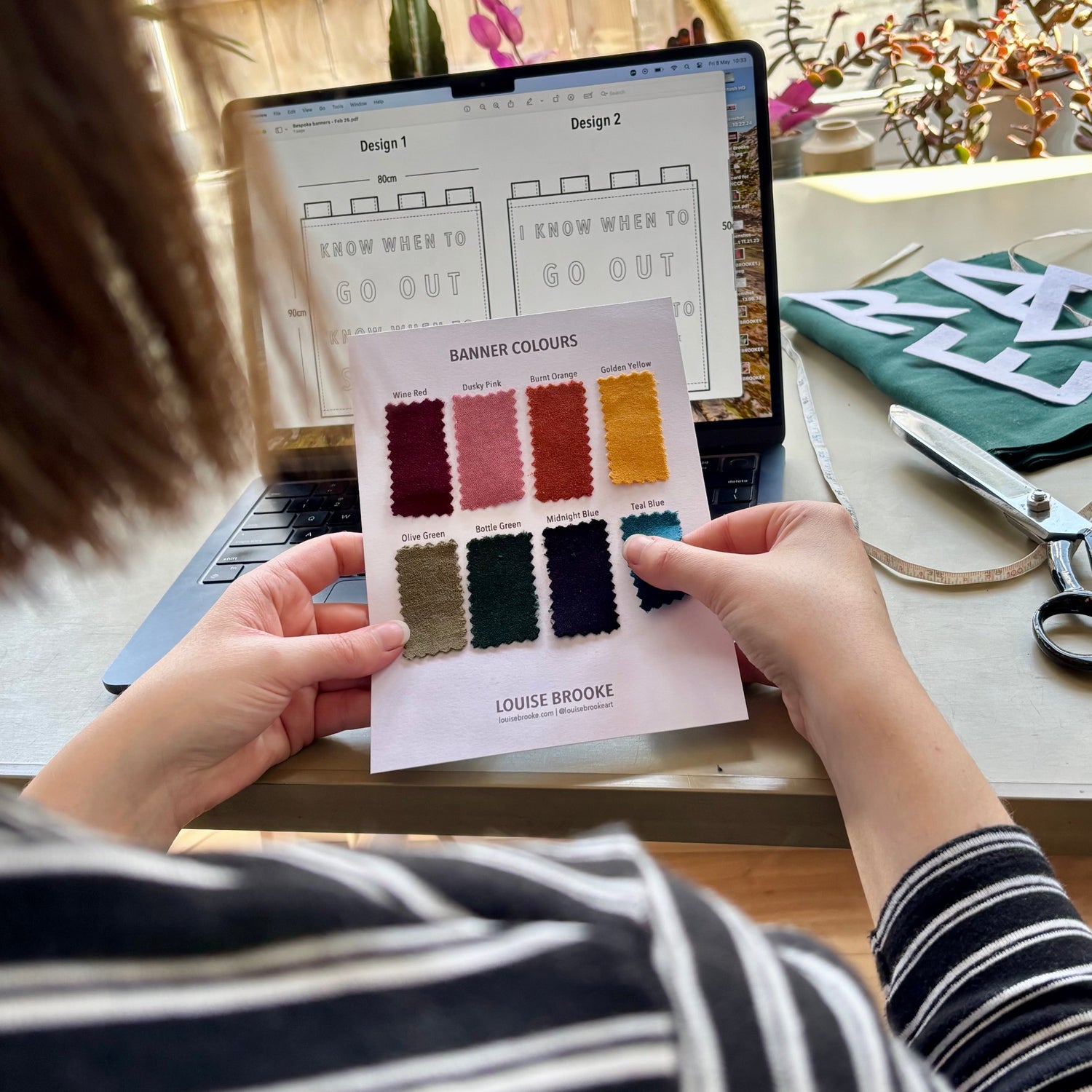 Person holding a color swatch card in front of a laptop displaying banner designs.