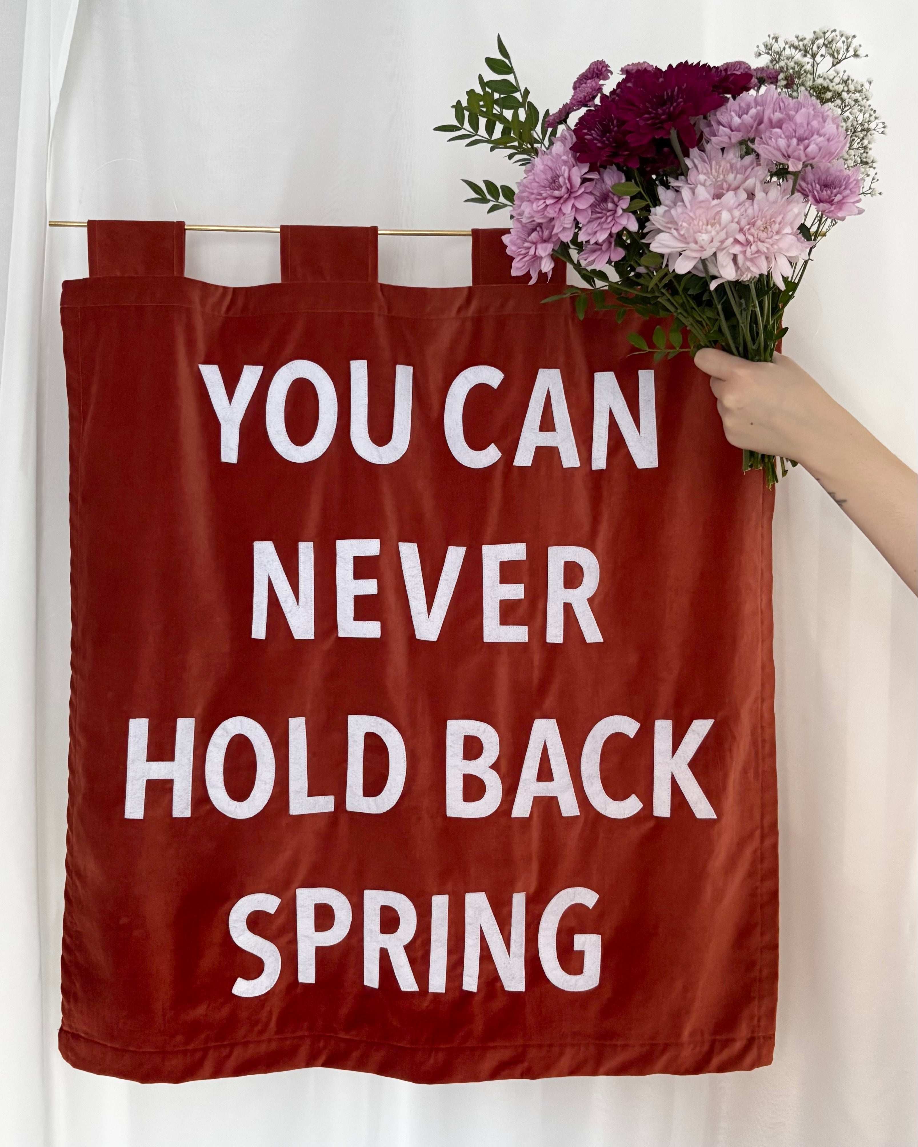 An orange velvet banner with "YOU CAN NEVER HOLD BACK SPRING" with a pink bouquet of flowers held up next to it.