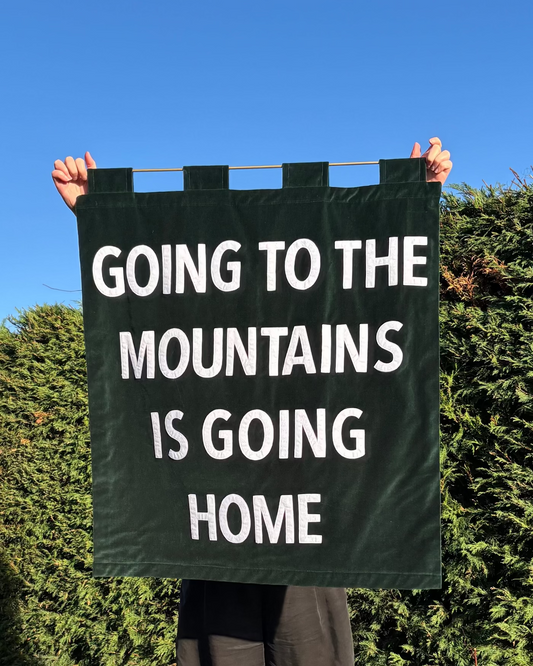 Person holding a deep green velvet banner with 'Going to the mountains is going home' text against a natural background.