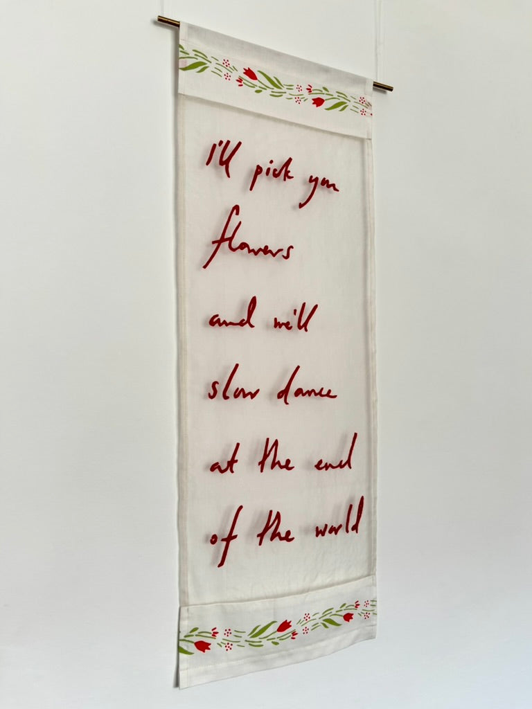 Light silk textile artwork which reads "I'll pick you flowers and we'll slow dance at the end of the world" in red handwriting, edged with floral fabric.