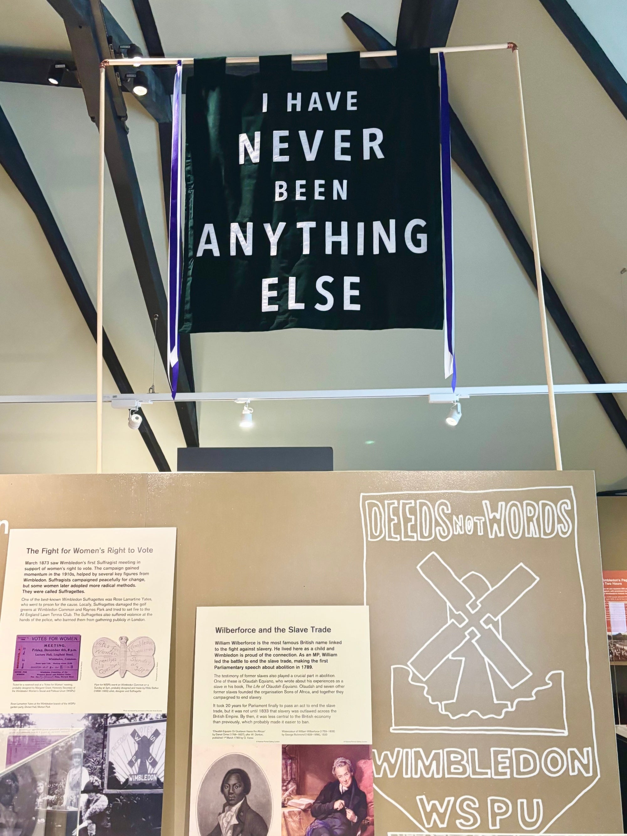 A green velvet banner with white text reading 'I HAVE NEVER BEEN ANYTHING ELSE' hangs from a white frame in an indoor exhibition space. Below, display boards show historical content including the Wimbleton and WSPU emblem.