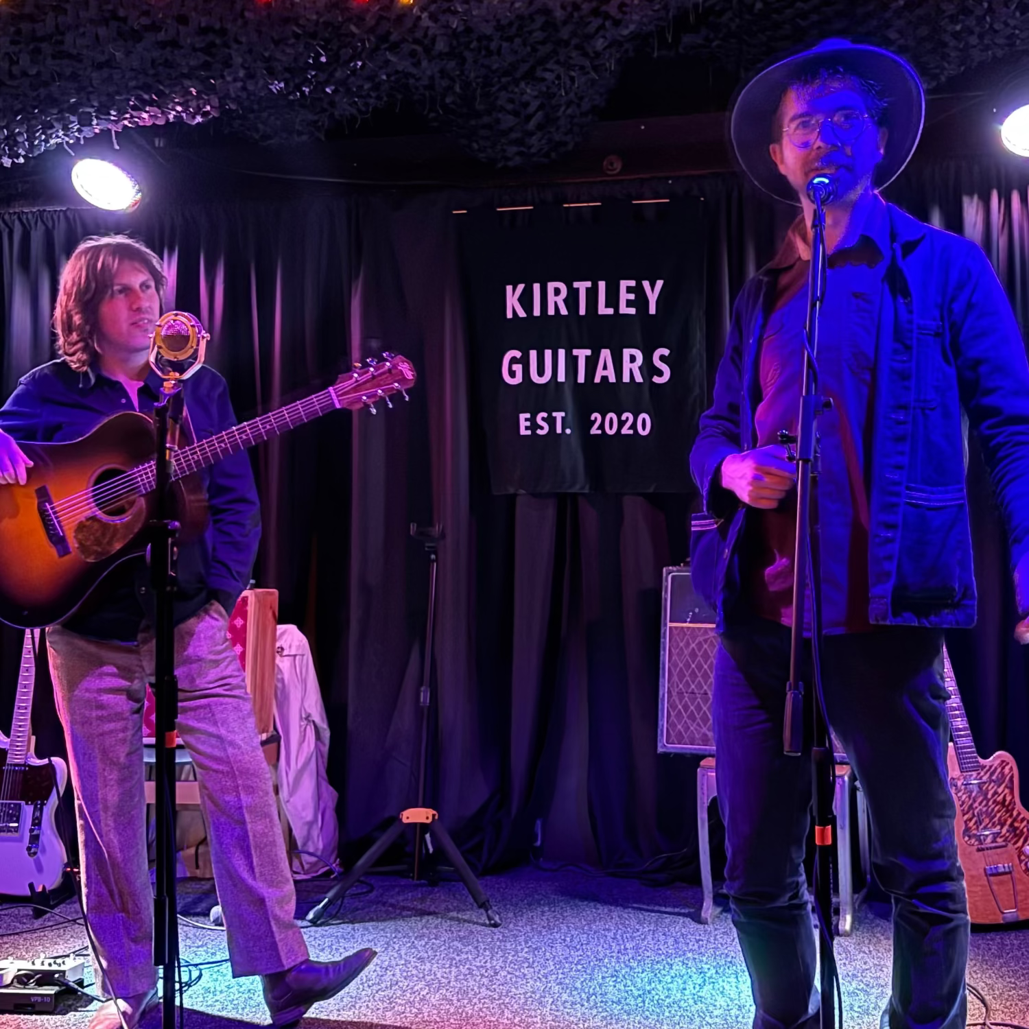 Two musicians performing on stage with 'Kirtley Guitars' banner in the background.