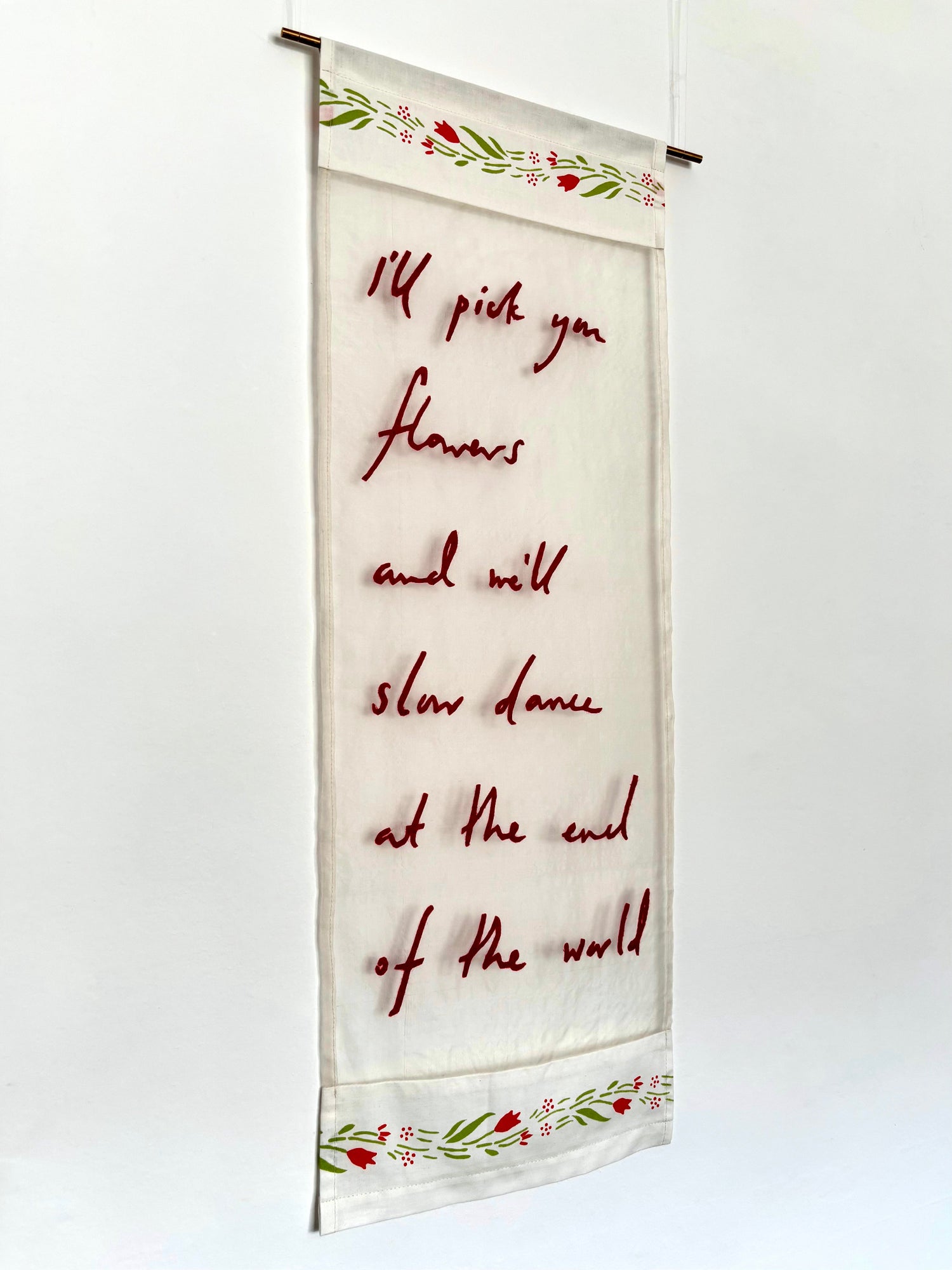 A vertical fabric wall hanging with a cream background, featuring red cursive text that reads, 'I'll pick you flowers and we'll slow dance at the end of the world,' with decorative red and green floral borders at the top and bottom.
