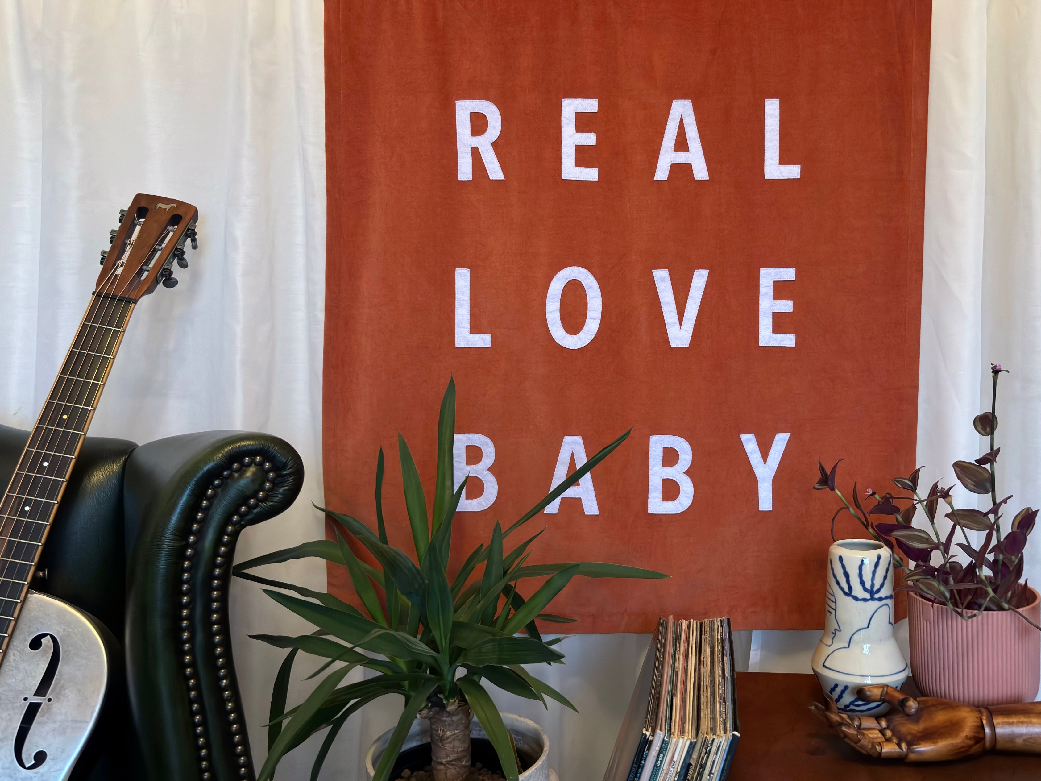 Burnt orange velvet banner with 'REAL LOVE BABY' text in a home setting with plants and a guitar.