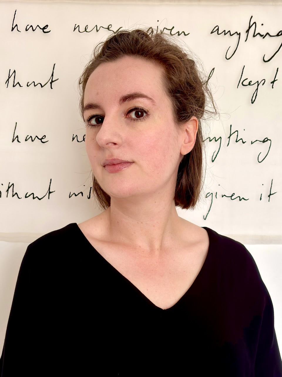 A woman with light brown hair tied back, wearing a black V-neck top, standing in front of a sheer textile artwork with handwritten black text.