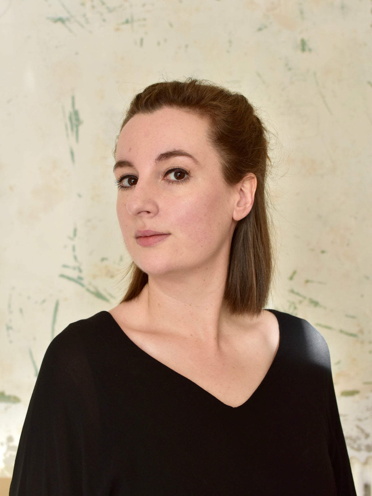 Artist Louise Brooke - a woman with brown hair pulled back, wearing a black V-neck top, photographed against a textured light beige wall.