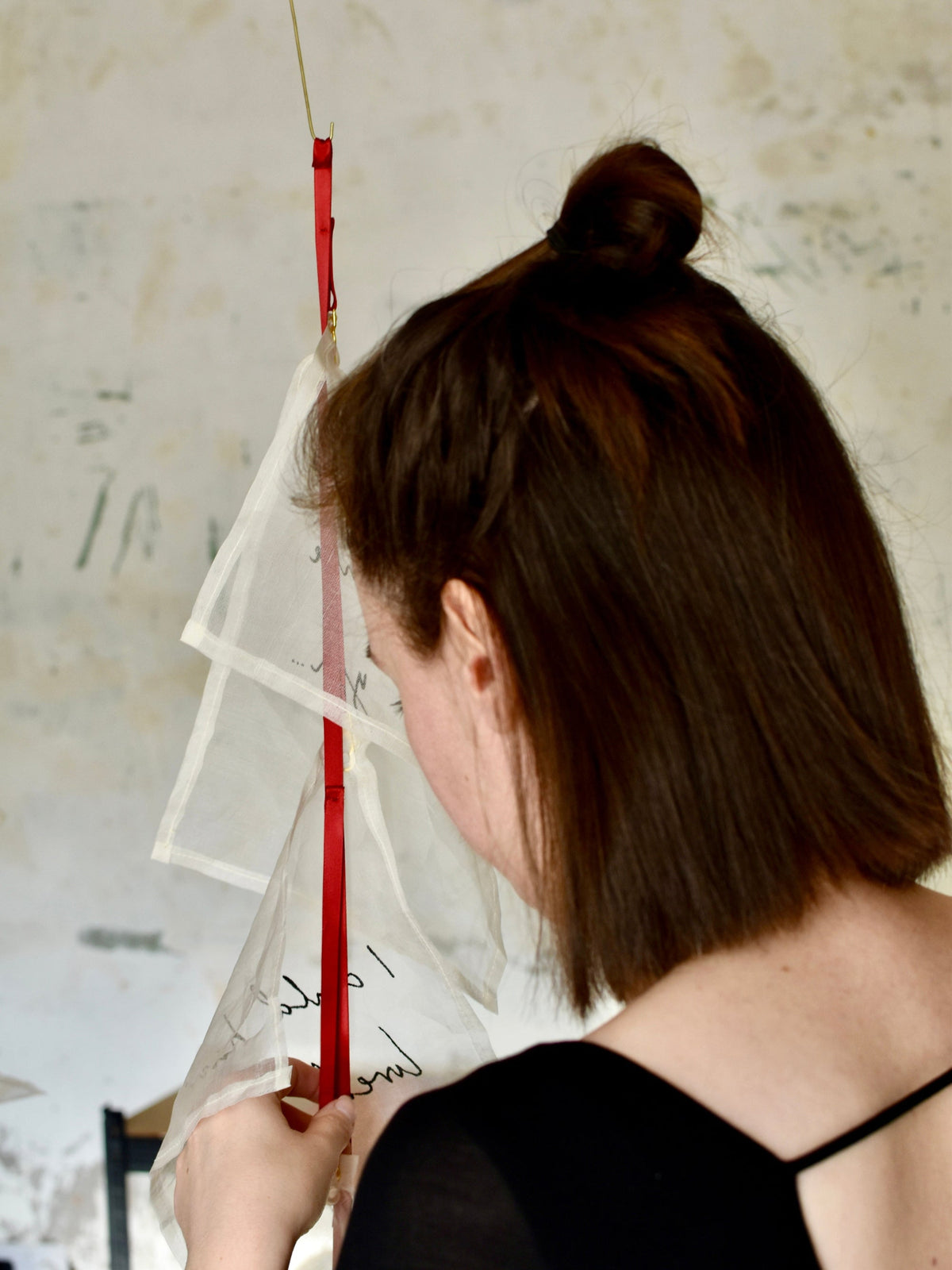 Artist Louise Brooke in her studio - a woman with brown hair in a bun is shown from behind, wearing a black top and holding a white translucent fabric panel with red ribbons against a textured off-white wall.