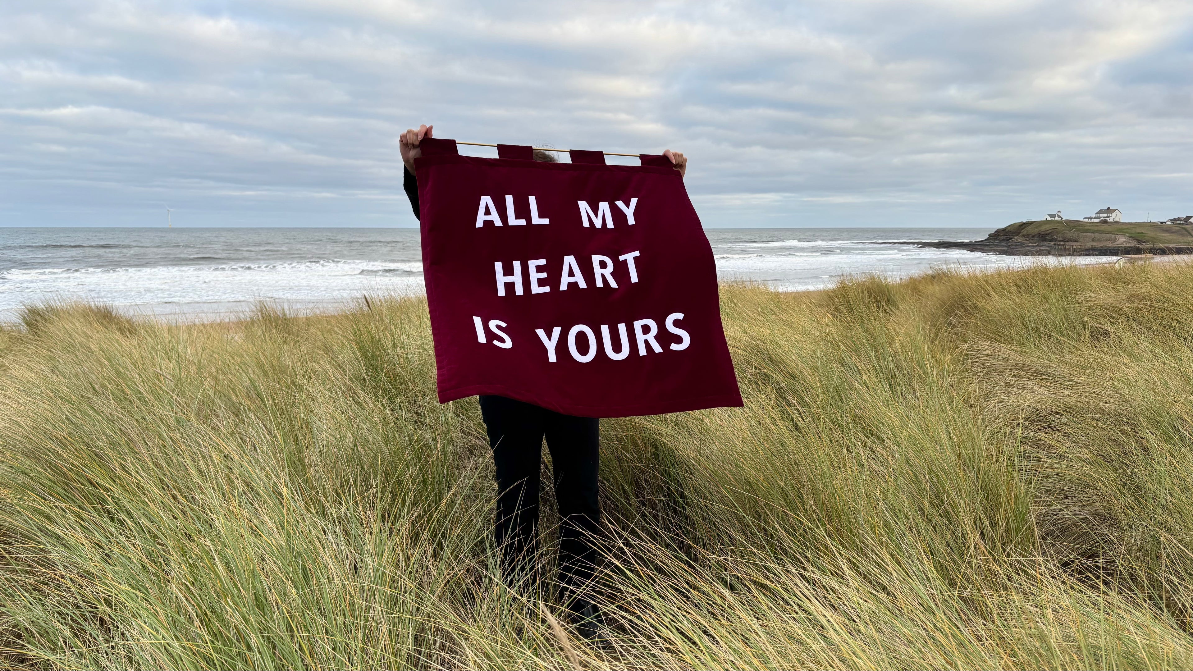 Person holding a red velvet banner with 'All my heart is yours' on a beach