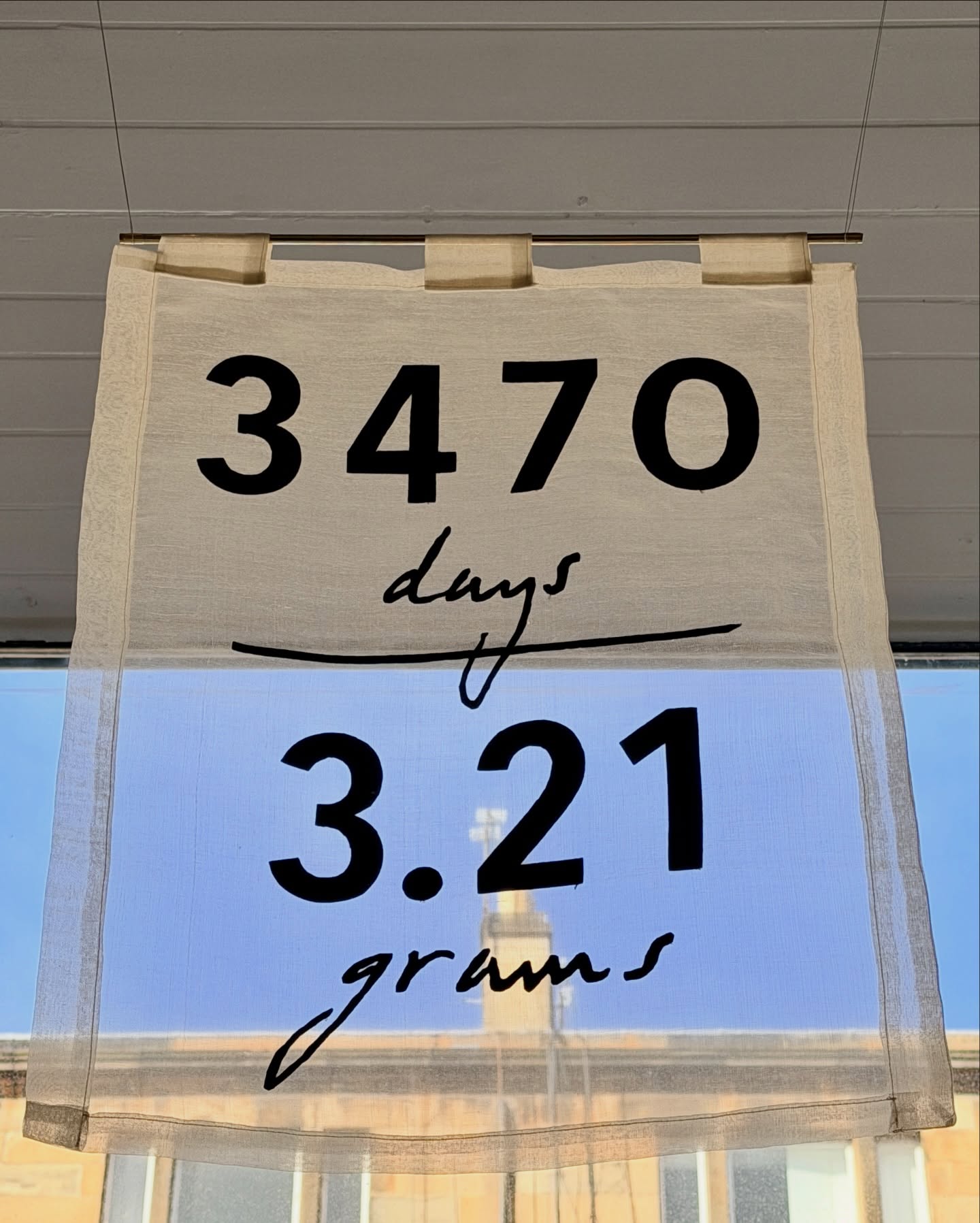 Fabric banner in an art gallery window with '3470 days 3.21 grams' text against a blurred outdoor background