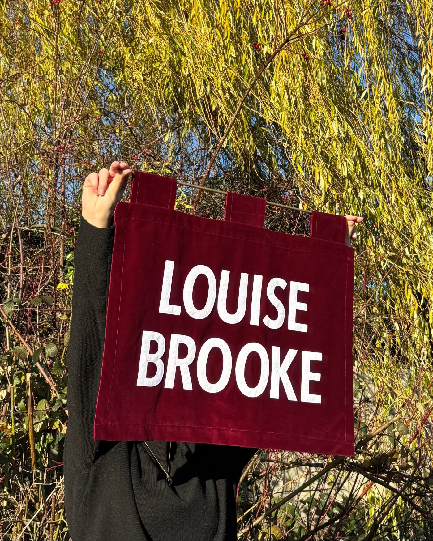 Person holding a red velvet banner with 'LOUISE BROOKE' text against a natural background