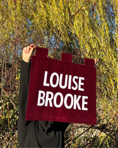 Person holding a red velvet banner with 'LOUISE BROOKE' text against a natural background