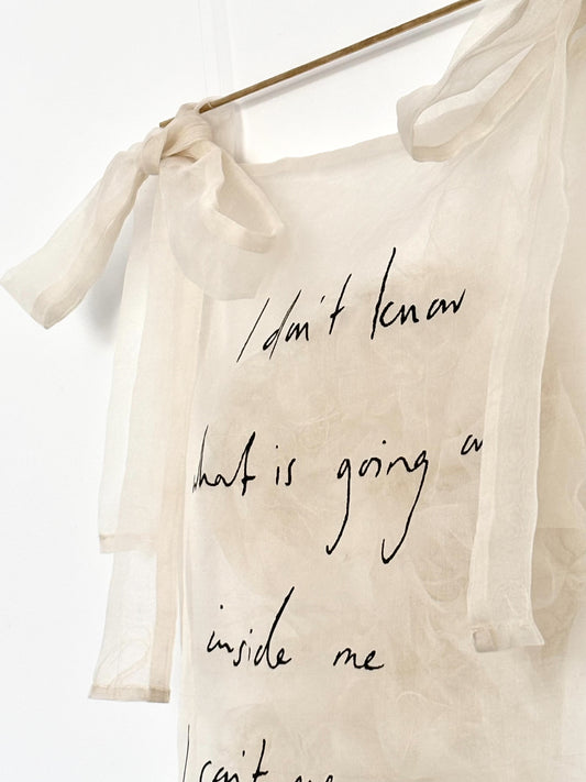 A sheer silk wall hanging with tied side bows and a brass pole, featuring handwritten black text that reads, 'I don't know what is going on inside me I can't even explain it to myself,' displayed against a white wall.