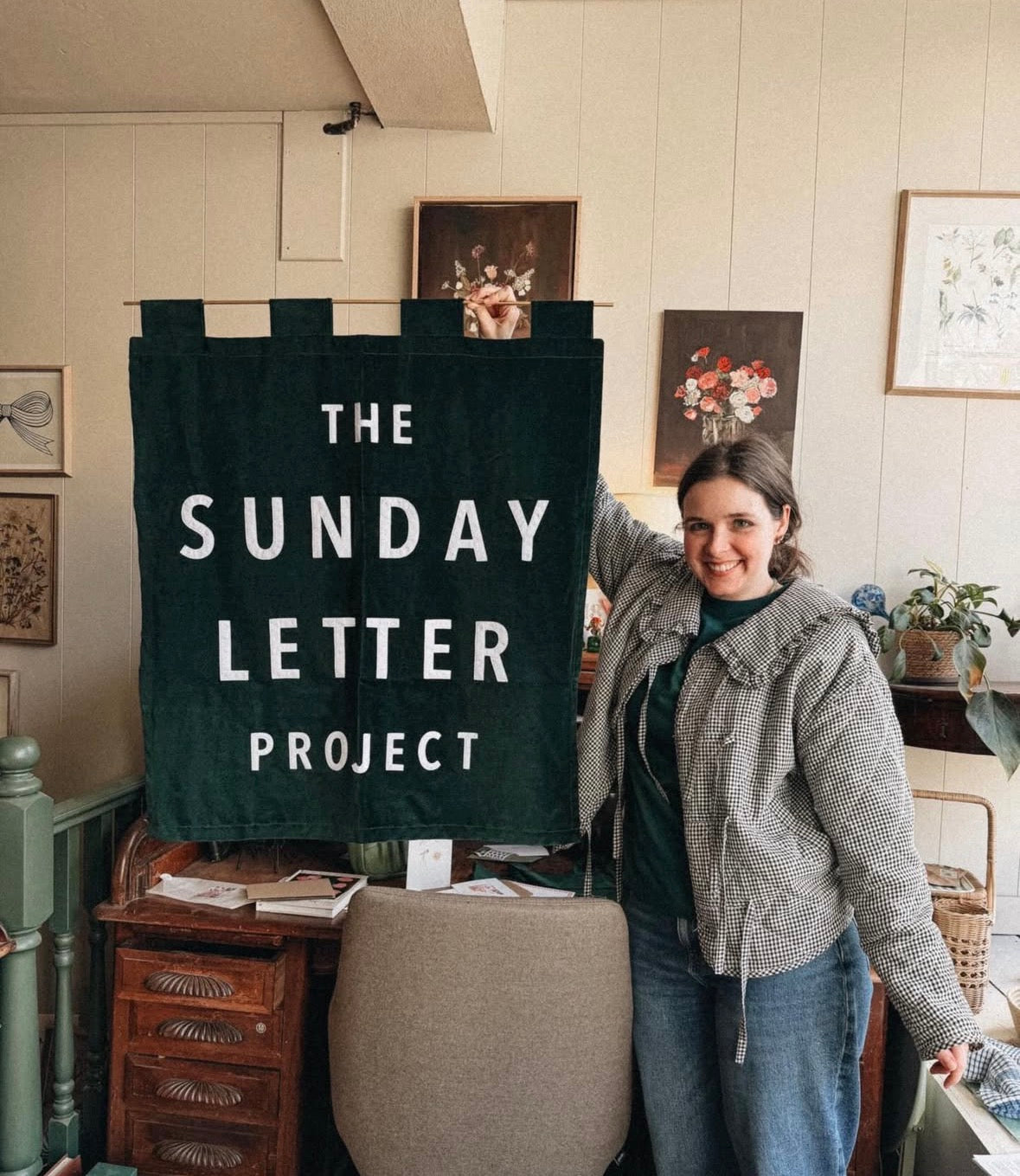 Person holding a banner with 'The Sunday Letter Project' in a room with framed pictures on the wall.