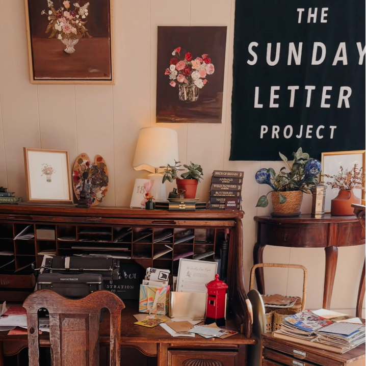 Vintage wooden desk and chair with letters against a wall with flower paintings and a green velvet banner which reads "The Sunday Letter Project"