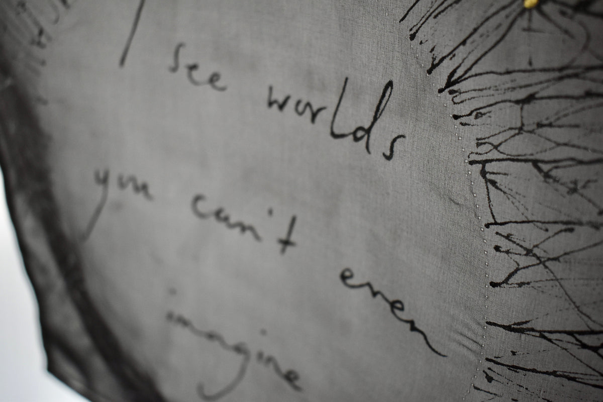 A close-up of black silk with black handwritten text reading 'I see worlds you can't even imagine' and black sketched lines on the right side.