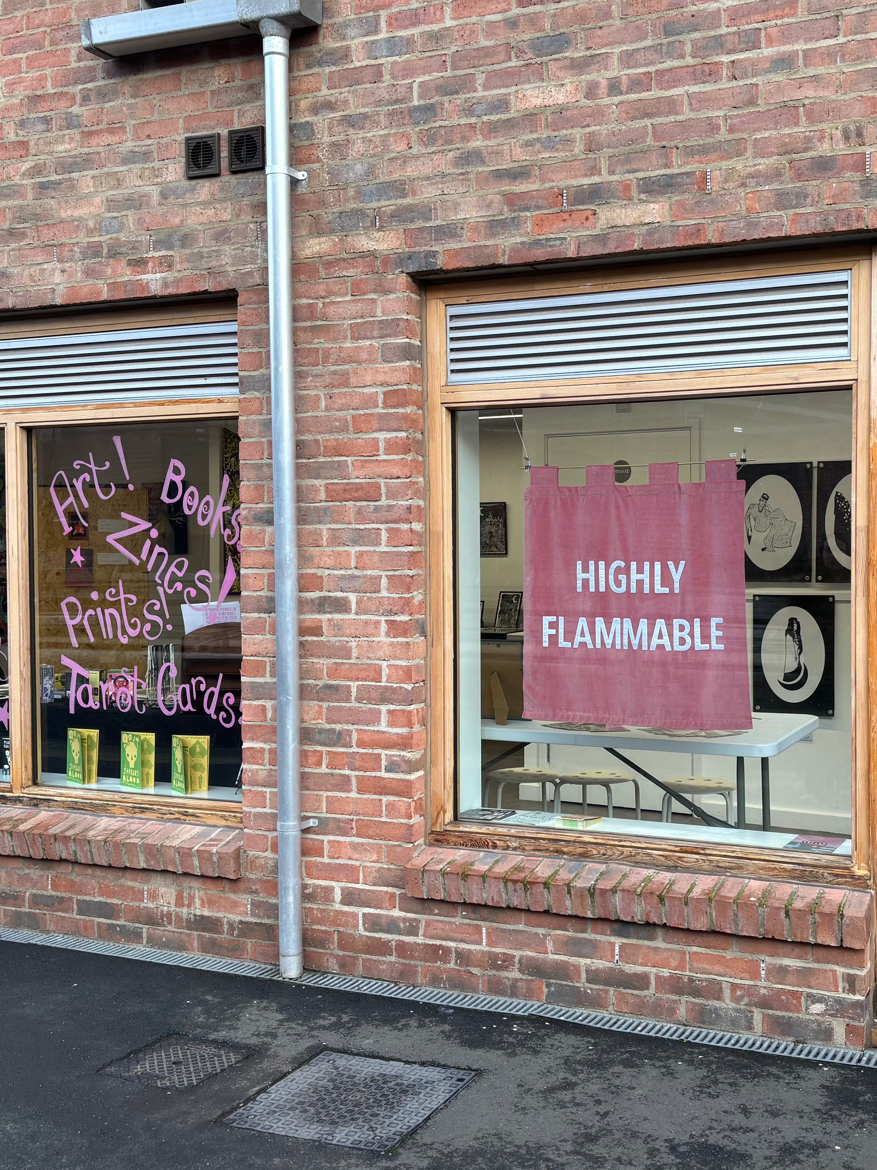 Brick building with two windows displaying signs, one of which is a pink velvet banner reading 'Highly Flammable'.