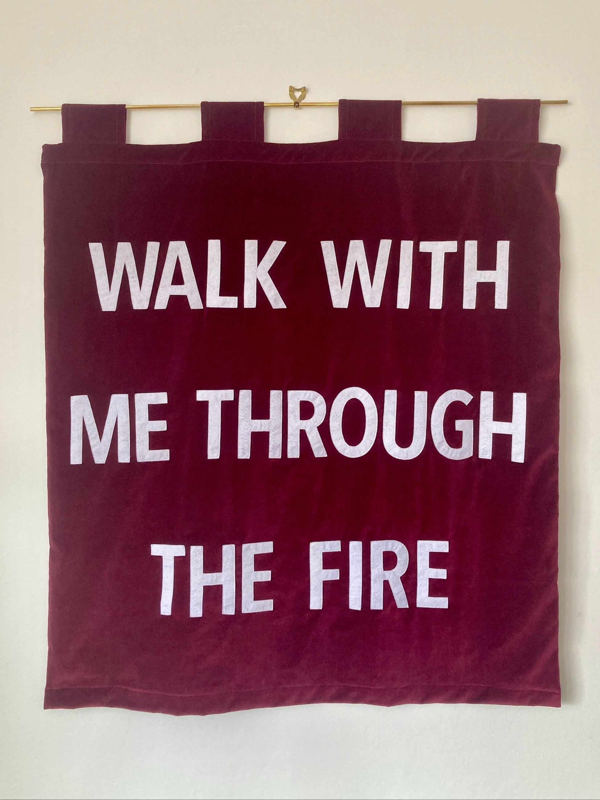 Red velvet banner with white text 'WALK WITH ME THROUGH THE FIRE' hanging on a brass rod against a white wall.