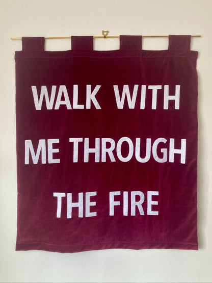 Red velvet banner with white text 'WALK WITH ME THROUGH THE FIRE' hanging on a brass rod against a white wall.