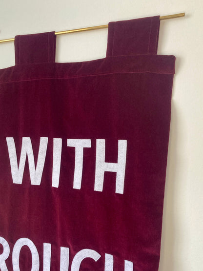 Close up of a red velvet banner with white text hanging on a brass rod against a white wall.