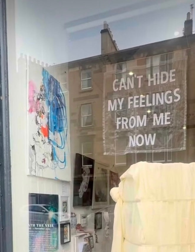Art gallery window display with paintings and sheer banner which reads 'CAN'T HIDE MY FEELINGS FROM ME NOW'.
